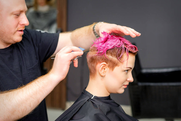 Стрижка фарбованого короткого рожевого вологого волосся молодої білої жінки чоловічим перукарем у перукарні
 - Фото, зображення