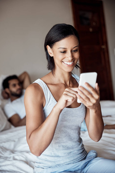 Mi sto svegliando con delle belle notizie. una giovane donna che usa il cellulare seduta su un letto con il marito sullo sfondo - Foto, immagini