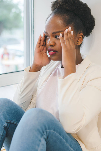 Mulher afro-americana sentada em casa perto da janela massageando templos sente dor de cabeça. Conceito de depressão. Saúde mental. - Foto, Imagem
