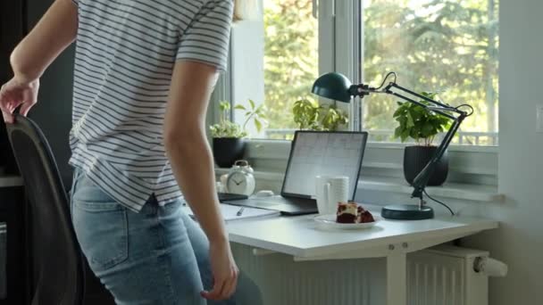 Woman sits down at desk near window and prepares to work. Home office workplace with laptop, notebook, cup of coffee and cake. Female designer working from home - Footage, Video