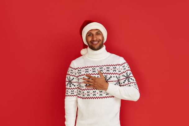Happy Man Smiling Camera. Cheerful Guy in Santa Hat Smiling Sincerely, Holding Hand at Chest, Positive Mood. Indoor Studio Shot, Isolated on Red Background  - Foto, Imagem