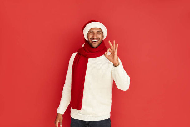 Smiling Man Showing Ok Gesture. Satisfied Multiracial Guy in Santa Hat Making Ok Symbol with Fingers, Approving, Satisfied. Indoor Studio Shot Isolated on Red Background  - Foto, Bild