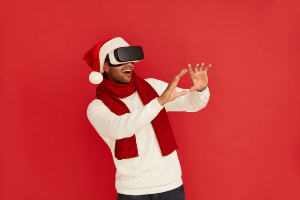 Focused Man Trying VR. Portrait of Multiracial Amazed Guy in Santa Hat Discovering New Technologies Wearing Virtual Reality Headset, Futuristic 3d Vision. Indoor Studio Shot Isolated Red Background  - Photo, image