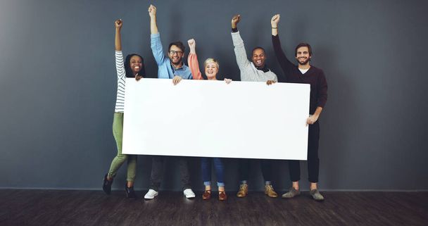 Say it loud, say it proud. Studio shot of a diverse group of people holding a blank placard and cheering against a gray background - Foto, imagen