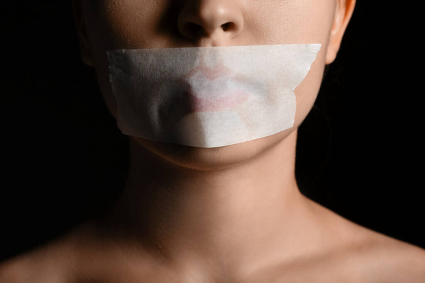 Νεαρή γυναίκα με κολλημένο στόμα σε σκούρο φόντο, κοντινό πλάνο. Έννοια λογοκρισίας - Φωτογραφία, εικόνα