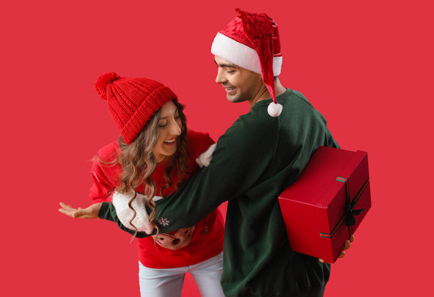 Young man surprising his beloved girlfriend with Christmas gift on red background - Foto, Imagem
