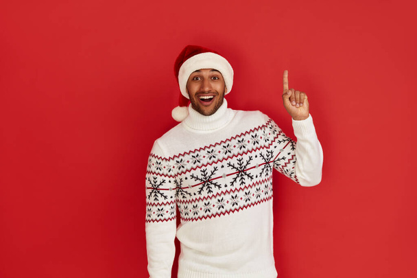 Shocked Man Pointing Aside. Astonished Multiracial Guy in Santa Claus Hat Pointing at Copy Space and Looking With Amazement. Indoor Studio Shot Isolated on Red Background  - Foto, imagen