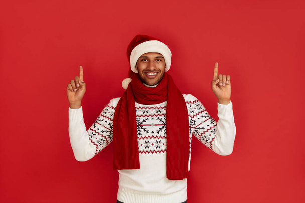 Smiling Man Pointing Hand. Positive Multiracial Guy Pointing Away Paying Your Attention at Empty Space for Advertisement. Indoor Studio Shot Isolated on Red Background  - 写真・画像