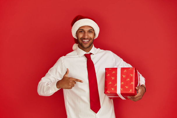 Man Pointing New Year Present. Multiracial Guy in Santa Hat Paying Attention at Gift Box and Looking at Camera. Red Background, Studio Shot  - 写真・画像