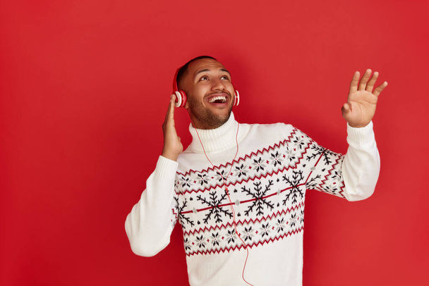 Winter Man Listening Music. Positive Multiracial Man Listening Favourite Song Wearing Headphones and Dancing. Indoor Studio Shot Isolated on Red Background  - 写真・画像