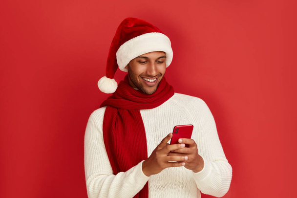 Happy Man met Smartphone. Multiraciale Guy in Santa Hat Lachen tijdens het gebruik van mobiele telefoon, chatten in sociale media, Genieten van mobiele service. Lege kopieerruimte voor reclame, Indoor Studio Shot  - Foto, afbeelding