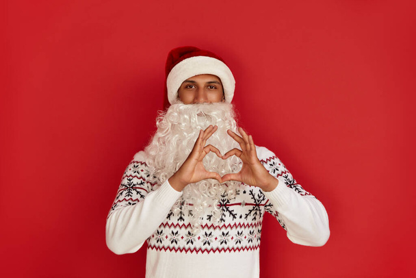 Santa Claus Showing Heart Shape Gesture. Positive Multiracial Man Holding Hands in Shape of Heart Showing Romantic Gesture, Love Confession, Valentines Day Celebration. Red Background  - Foto, Bild