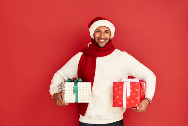 Smiling Man Holding Presents. Positive Multiracial Guy in Santa Claus New Year Hat Holding Gift Boxes, Delivery of Holiday Presents. Indoor Studio Shot Isolated on Red Background  - Foto, immagini