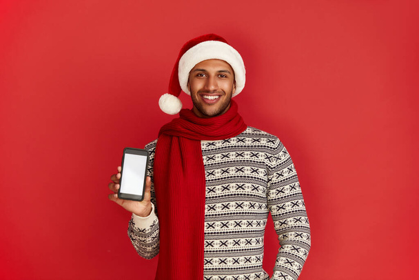 Happy Man Using Smartphone. Multiracial Guy in Santa Hat Smiling while Showing Cellphone, Chatting in Social Media, Enjoying Mobile Service. Empty Copy Space for Advertisement, Indoor Studio Shot  - Foto, afbeelding