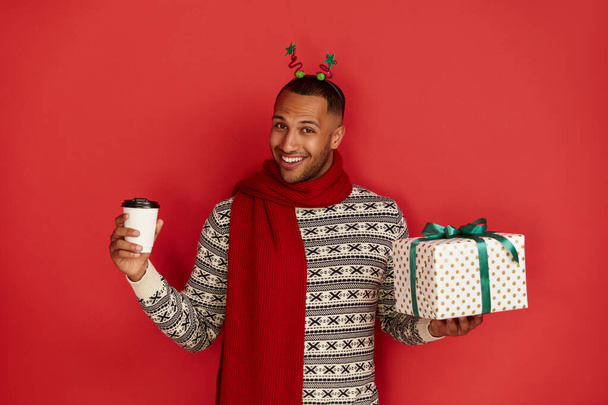 Happy Man Holding Present. Excited Multiracial Guy Holding in Coffee to Go and Wrapped Present Box with Excitement. New Year Concept. Indoor Studio Shot Red Background  - Foto, immagini