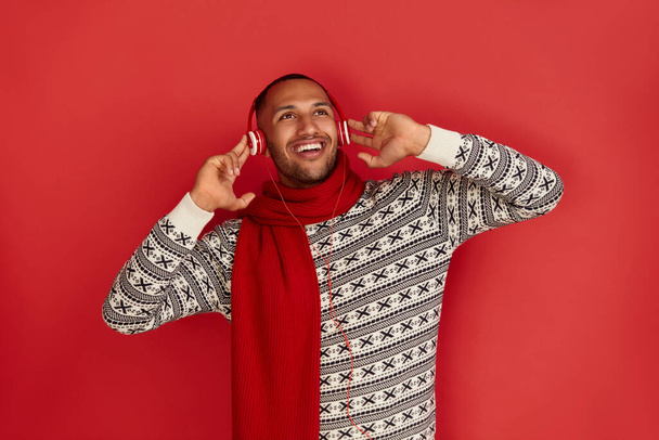 Winter Man Listening Music. Positive Multiracial Man Listening Favourite Song Wearing Headphones and Dancing. Indoor Studio Shot Isolated on Red Background  - Zdjęcie, obraz
