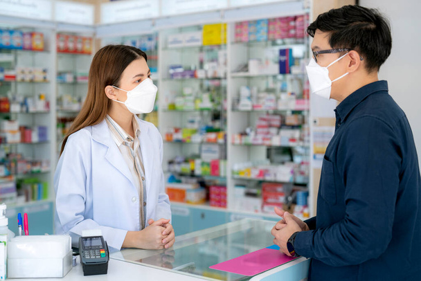 Aasialainen apteekkarinainen, jolla on suojanaamari apteekissa myymälässä myytävänä lääke ja apteekki asiakkaansa kanssa  - Valokuva, kuva
