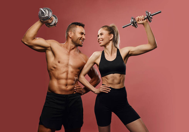 Sport handsome couple with dumbbells at pink background. Fitness male and female models looking each other in studio. Happy gym partners with sport equipment. Shirtless man and sexy girl with six pack - Foto, Imagen