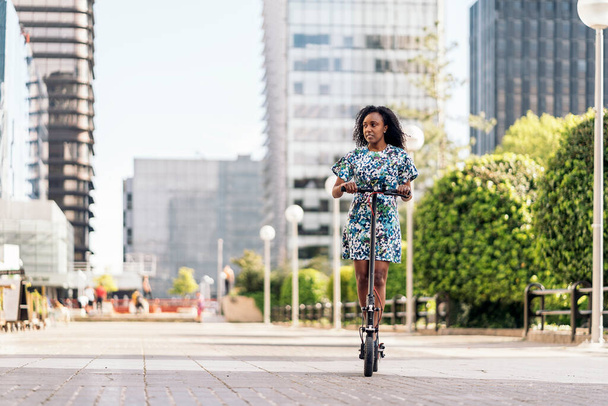 Young black woman with curly hair having fun in the city riding her electric scooter. - 写真・画像