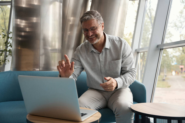 Uomo sorridente in occhiali sta comunicando attivamente online con un collega, è seduto su un divano blu - Foto, immagini