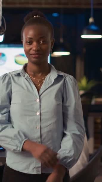 Vídeo vertical: Mulher de negócios afro-americana sorridente cruzando retrato de armas, visão frontal tiro médio. Empregado da empresa em pé na porta da diretoria no espaço de co-trabalho, apresentação de colegas de trabalho em - Filmagem, Vídeo