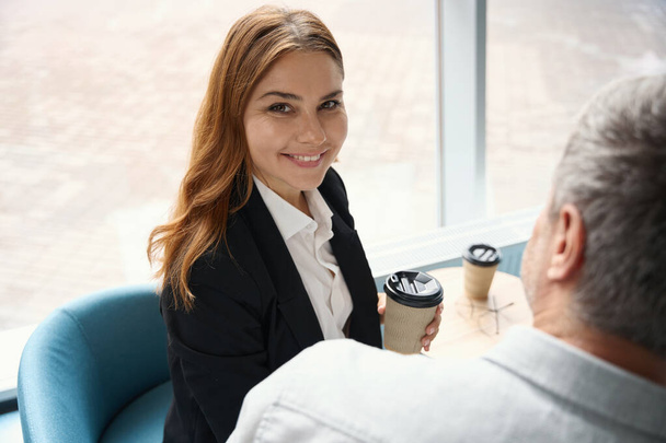Χαρούμενη κοκκινομάλλα γυναίκα επικοινωνεί με έναν άνδρα συνάδελφο που βρίσκονται με καφέ κοντά στο παράθυρο - Φωτογραφία, εικόνα