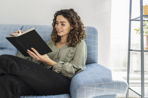 Lächelnde junge kaukasische Frau mit lockigem Haar liest auf dem Sofa ein Buch. - Foto, Bild