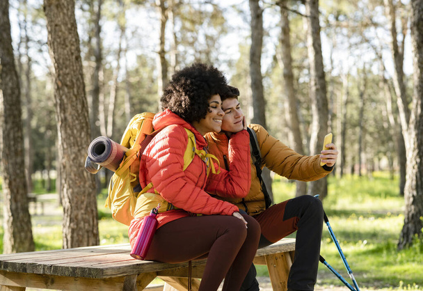 Multiraciální pár v lese pěší turistiku a brát selfie s chytrým telefonem sedí na piknik prostoru. Afričan americká žena a kavkazský muž - Fotografie, Obrázek