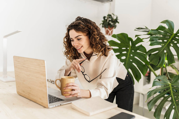 Junge Frau mit lockigem Haar trinkt Kaffee, während sie auf den Laptop schaut und zu Hause eine Brille hält - Foto, Bild