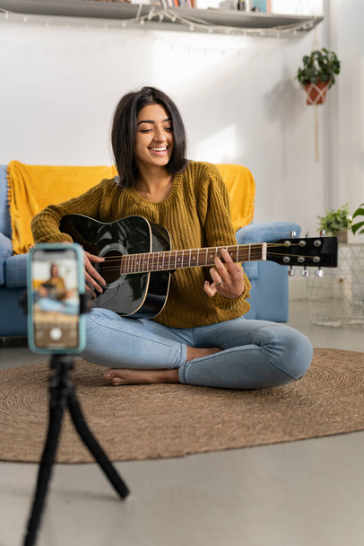 Młoda brunetka indianka grająca na gitarze akustycznej podczas nagrywania filmiku ze smartfonem. Tysiącletnia Hinduska kobieta w domu. - Zdjęcie, obraz