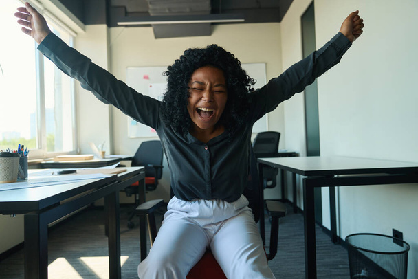 Emotional young african american woman in white trousers and gray shirt sits in office chair, laughs with a victory gesture - Fotoğraf, Görsel