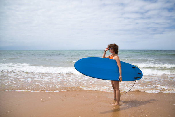 Attractive mature woman with curly hair, sunglasses and bikini, posing holding a blue surfboard under her arm. Concept sea, sand, sun, beach, vacation, surf, summer. - 写真・画像