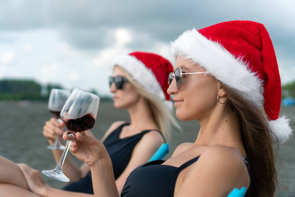 Portret van twee jonge vrouwen in badpakken, zonnebril en kerstmanhoeden met een glas wijn in hun handen zittend op klapstoelen op het zandstrand van de zee en genietend van het moment. - Foto, afbeelding