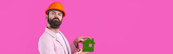 Ritratto di giovane ingegnere edile indossare cappello rigido arancione, in una giacca rosa in piedi su sfondo rosso studio. Un uomo ha una eco-casa verde. - Foto, immagini