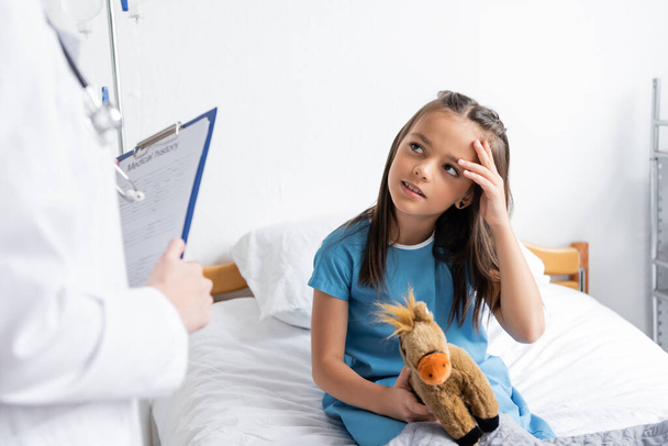 Girl holding soft toy and touching head near blurred pediatrician with clipboard in clinic  - Photo, Image