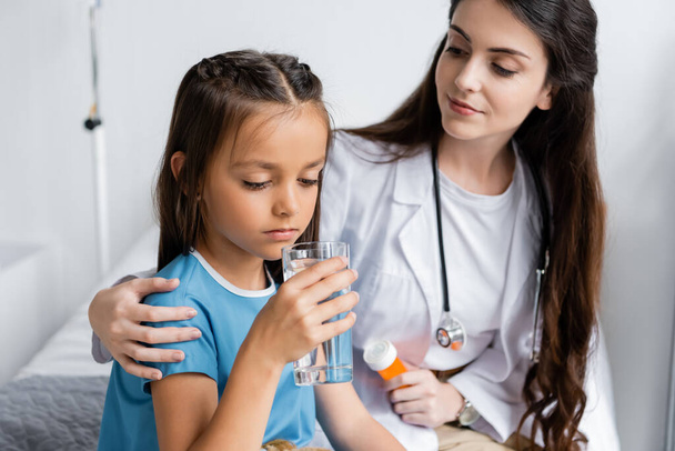 Pédiatre tenant des pilules près de l'enfant avec un verre d'eau dans la salle d'hôpital  - Photo, image