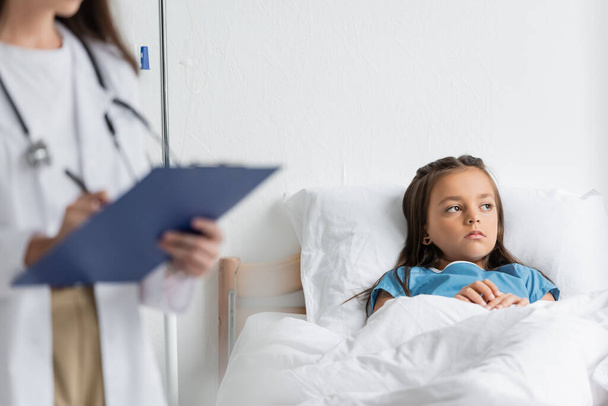 Niño triste mirando hacia otro lado mientras está acostado en la cama cerca de un médico borroso con portapapeles  - Foto, Imagen