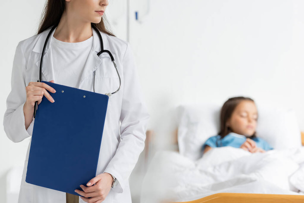 Pediatrician holding clipboard near blurred patient in hospital ward  - Фото, изображение