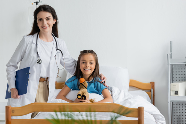 Médecin souriant étreignant enfant heureux avec jouet doux dans la salle d'hôpital  - Photo, image