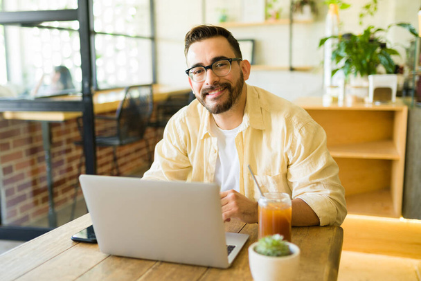 Attractive happy man smiling at the coffee shop while working with his laptop  - Photo, Image