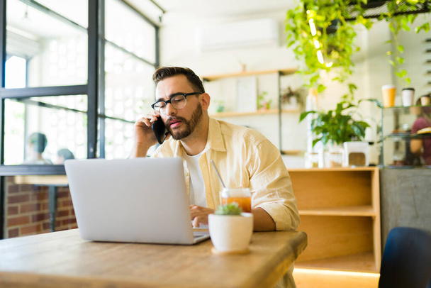Ammattimainen mies puhuu puhelimessa asiakkaan kanssa työskennellessään kahvilan kannettavan tietokoneen kanssa - Valokuva, kuva
