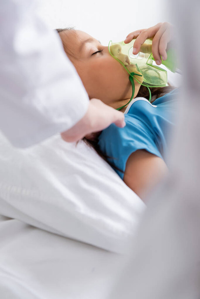 Wazig kinderarts aanraken kind met zuurstofmasker op bed in kliniek  - Foto, afbeelding