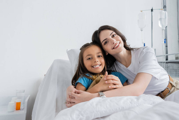 Θετική γυναίκα και κόρη κοιτάζοντας κάμερα στο κρεβάτι στο θάλαμο του νοσοκομείου  - Φωτογραφία, εικόνα