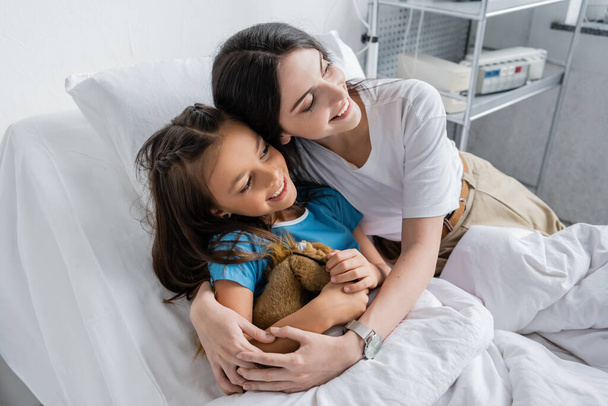 Happy parent hugging daughter with toy on bed in clinic  - Foto, Imagem