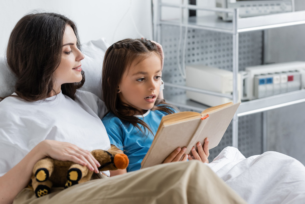 Kid reading book near mom with soft toy on hospital bed  - Фото, зображення