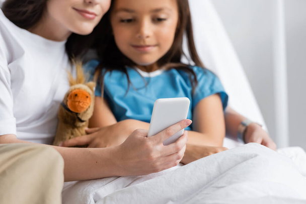 Mujer usando teléfono inteligente cerca de hija con juguete suave en la cama en el hospital  - Foto, imagen