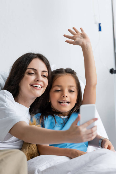 Cheerful woman holding smartphone near daughter during video call in clinic  - Photo, Image