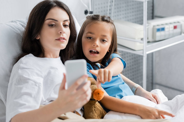 Ребенок указывает на смартфон рядом с мамой и мягкую игрушку на кровати в клинике  - Фото, изображение