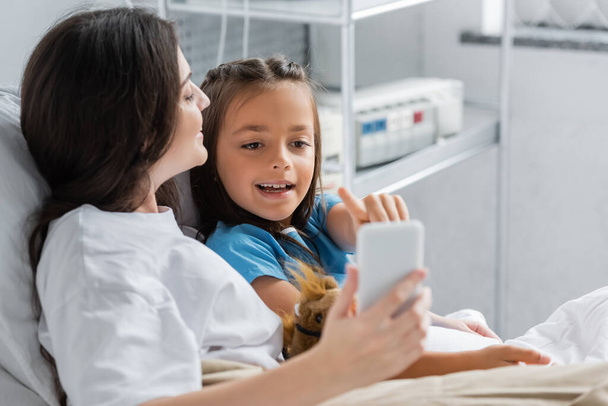 Menina sorridente apontando para o smartphone perto da mãe na cama na clínica  - Foto, Imagem