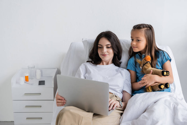 Mãe segurando laptop perto da filha em vestido de paciente na enfermaria  - Foto, Imagem